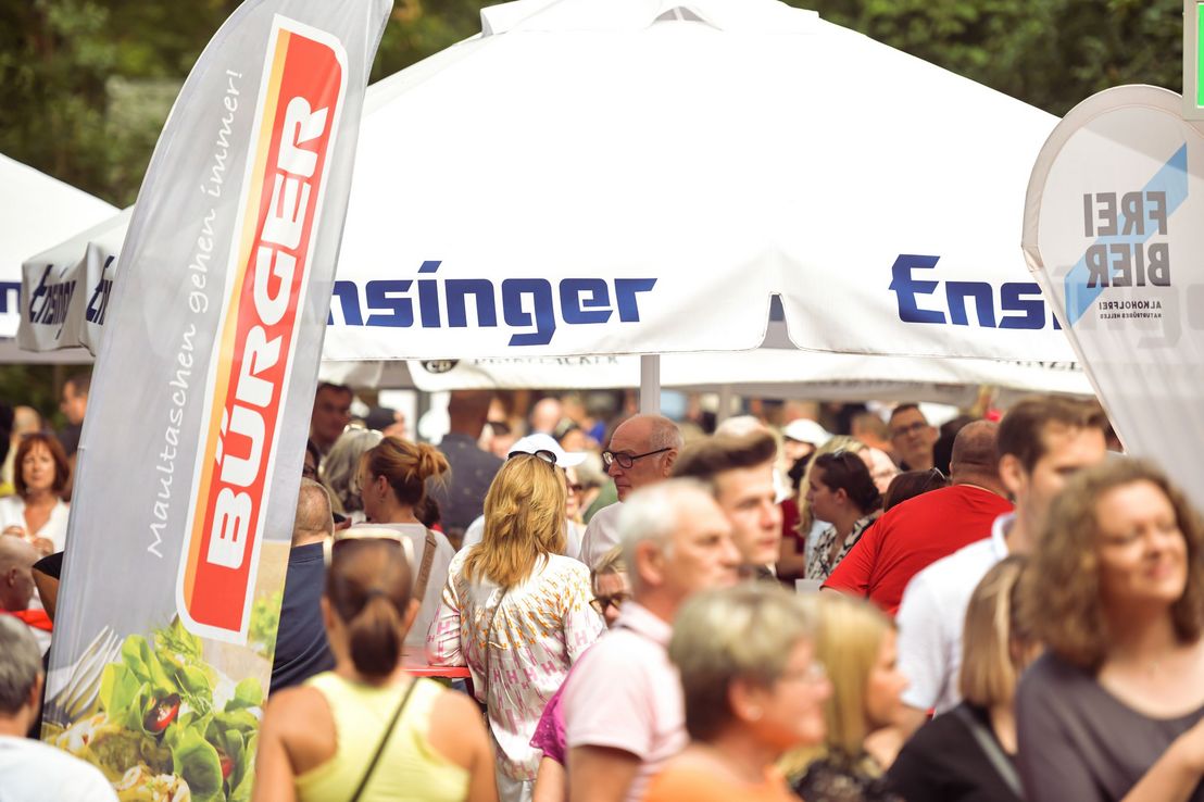 Menschen stehen an den Gastronomiehütten der BÜRGER Freilichtbühne unter großen Sonnenschirmen