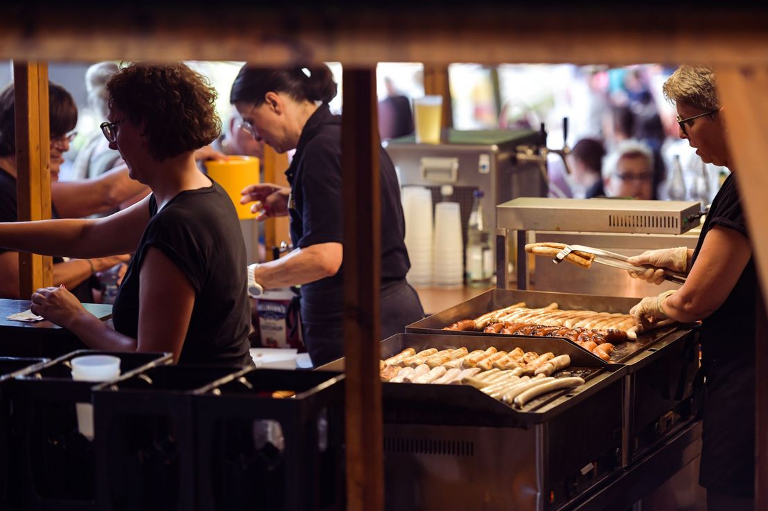 Mitarbeiter eines Gastronomie-Standes bereiten die Bestellungen der Besucher:innen zu